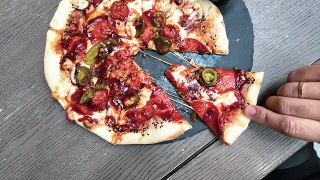 A close-up shot shows a hand lifting a cheesy pepperoni jalape&ntilde;o pizza slice from a rustic round pizza on a wooden table, with melting cheese and vibrant toppings in natural daylight.