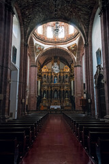 Obraz premium Grand cathedral interior with tall arches and golden altar. Dark pews line a red-laid floor leading to sacred space. Chandelier hangs above, casting soft glow