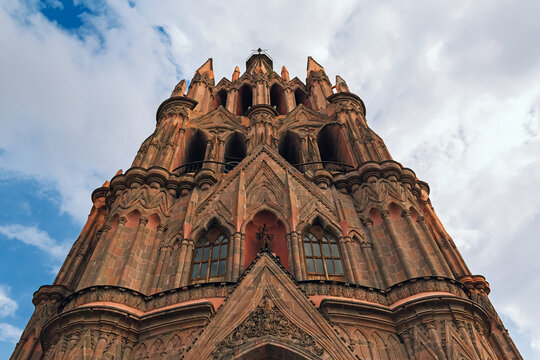 The imposing stone church of San Miguel Arcangel rises toward cloudy sky. Gothic spires frame a grand arched entrance. Palm trees flank a solemn, quiet cathedral. Iron gates and red pavement frame the