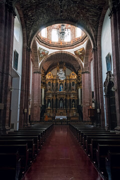 Grand cathedral interior with tall arches and golden altar. Dark pews line a red-laid floor leading to sacred space. Chandelier hangs above, casting soft glow