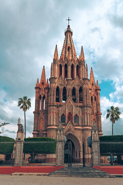 The imposing stone church of San Miguel Arcangel rises toward cloudy sky. Gothic spires frame a grand arched entrance. Palm trees flank a solemn, quiet cathedral. Iron gates and red pavement frame the
