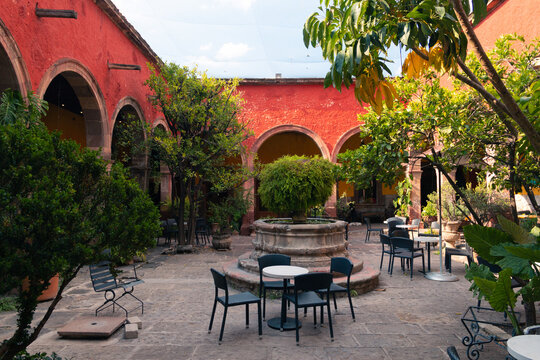 Red arches frame a tranquil courtyard. Stone table and chairs invite quiet conversation. Lush green trees soften the vibrant walls. Sunlight glows warmly on the cobblestone floor