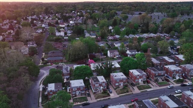 Sunset aerial drone view of a Philadelphia suburban neighborhood featuring brick twin homes, tree-lined streets, yards, and lush spring foliage at golden hour.
