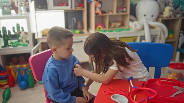 Girl uses toy stethoscope and syringe on seated boy at a preschool play table in building; playful roleplay learning.