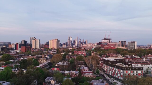 Aerial drone view of West Philadelphia rowhouses and tree canopy stretching toward Philadelphia's downtown skyline at dusk with active construction cranes.