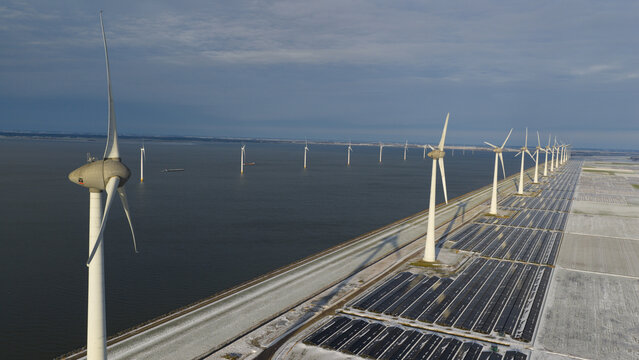 Aerial view of modern wind turbines and rows of solar panels along a coastal dike under a cloudy winter sky in Espel, Flevoland, Netherlands.