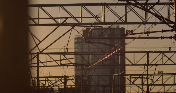 Dark silhouettes of railway catenary gantries and tangled power lines stand against a hazy orange sunset over a city skyline.