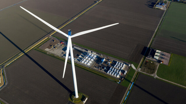 Aerial view of a large white wind turbine standing in flat agricultural fields with a battery storage facility at its base near Dronten, Flevoland, Netherlands.