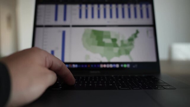 Close-up view of a laptop displaying a business analytics dashboard with statistics, pie charts, and country map. Hands type on the keyboard while using the mouse to click and scrolling.