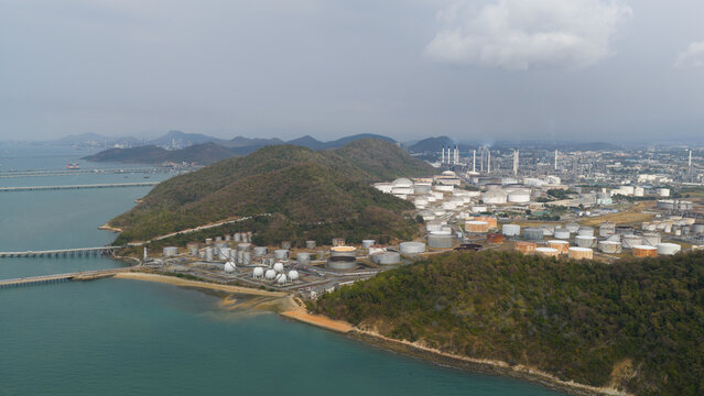 Aerial view of the Bangchak Sriracha Refinery featuring industrial storage tanks and chimneys along the coastline with lush hills in Si Racha District, Chon Buri, Thailand.