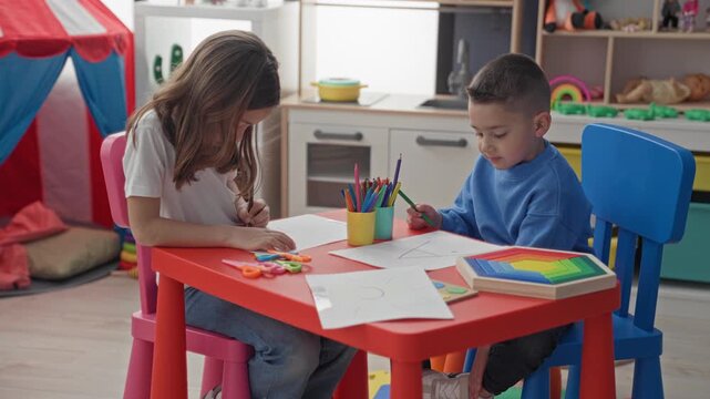 Girl and boy drawing with colored pencils at a red kids table, holding pencils and pointing to paper in a playroom; creative concentration.