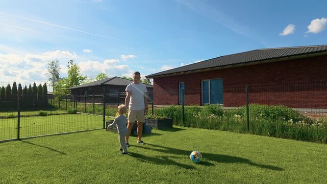 A dad and young boy play with a ball and frisbee in a fenced backyard on a sunny afternoon. Vibrant green grass, casual attire, clear blue sky, and soft natural lighting create a cheerful, family-frie