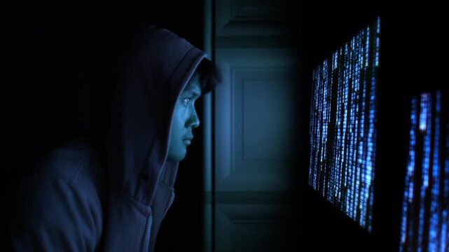 Hooded hacker illuminated by blue computer code screen in dark room, prepares to enter and work