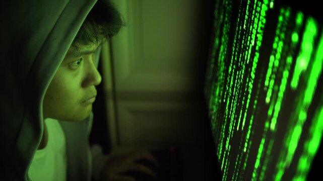 Hooded hacker using computer with glowing green code screen at night, hoodie up tapping and typing