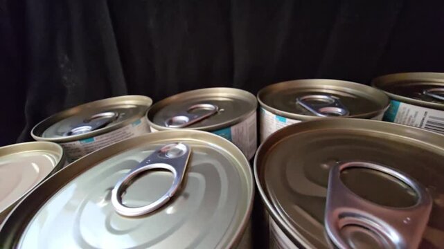 Close-up of multiple metal tin cans with easy-open ring pull tabs arranged against a dark background, ideal for food supply concepts.