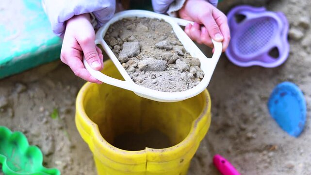 Experience the magic of childhood in this delightful scene where little hands enthusiastically sift and pour sand into a bucket. The carefree spirit of play permeates every moment.