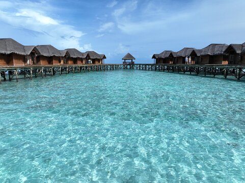 Aerial view of luxury overwater wooden villas with thatched roofs built on stilts over crystal clear turquoise lagoon water under a blue sky in Maldives.