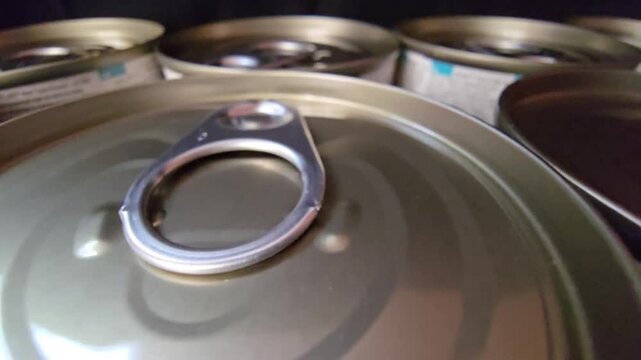 Close-up of multiple metal tin cans with easy-open ring pull tabs arranged against a dark background, ideal for food supply concepts.