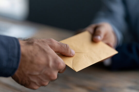 Two hands exchange a brown envelope. One hand holds it steady, the other offers it forward. Fingers show signs of wear, suggesting routine use. The envelope bears a subtle crease