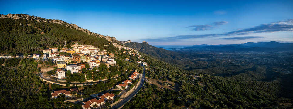 Aerial view of the mountain village nestled on a steep hillside with historic houses, dense green forests and winding roads overlooking a wide valley in Baunei, Sardinia, Italy.