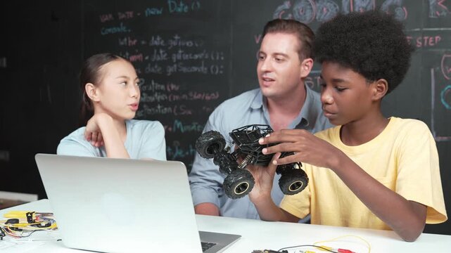 African boy inspect and fix car model with laptop and wire placed on table while teacher explain about robotic construction at blackboard with code or prompt at STEM class. Education. Edification