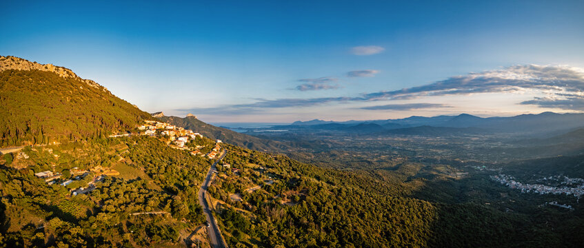 Aerial view of the mountain village of Baunei perched on a steep hillside with lush green slopes, a winding road, and a distant view of the coast Baunei, Sardinia, Italy.