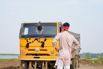 Indian truck driver standing confidently from the back beside a yellow dumper truck on a rural construction road © Niks Ads