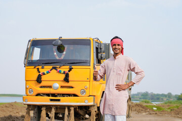 Smiling Indian truck driver posing proudly beside a yellow dumper truck on a rural roadside © Niks Ads