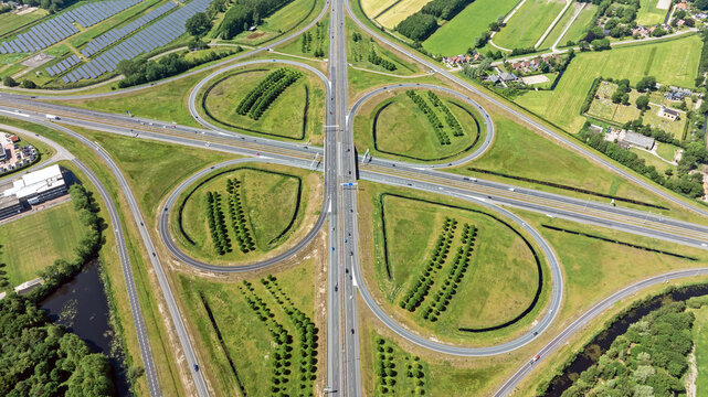Aerial view of a cloverleaf highway interchange with green fields, solar panels, and surrounding vegetation in Heerenveen, Friesland, Netherlands.