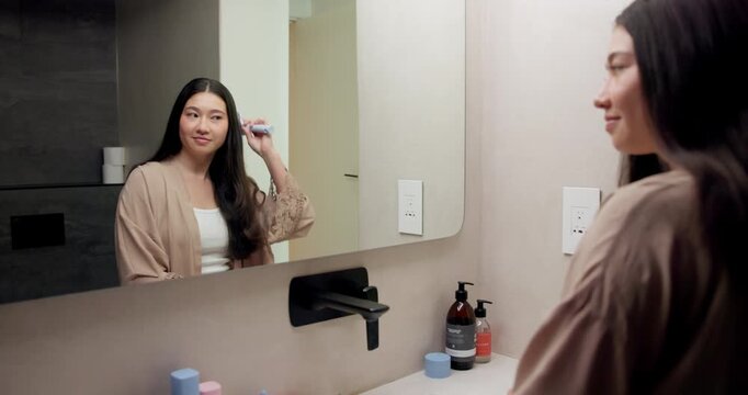 Mirror, beauty and woman brushing hair in bathroom for grooming, self care or morning routine. Happy, reflection and Asian female person with hairstyle for daily haircare treatment in apartment.