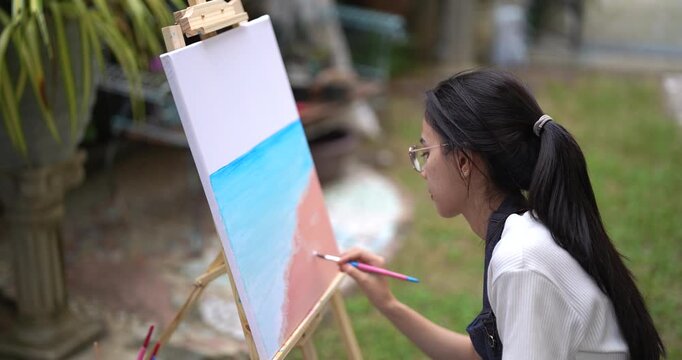 asian female artist painting beach scene with warm tones on canvas in backyard garden using brush carefully enjoying quiet hobby time during calm afternoon surrounded by soft light and nature