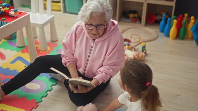 Woman reads book aloud, hand raised in gesture while seated with child on playroom floor; intergenerational warmth.