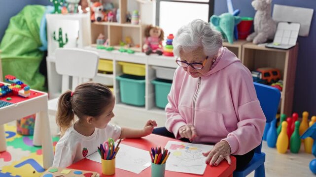 Woman and little girl sit at red table with paper and colored pencils, woman leans over and points to drawing in studio; gentle guidance warmth.