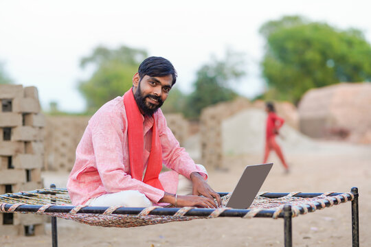 A rural entrepreneur in traditional attire sits on a charpai using a laptop at a brickwork site, giving a thumbs up
