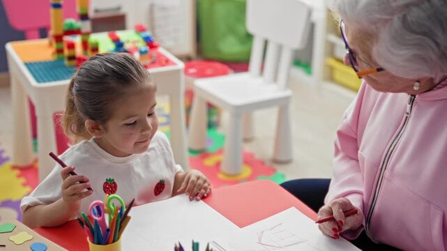 Woman guiding a little girl drawing with a pencil at a preschool table inside a classroom building; learning nurture.