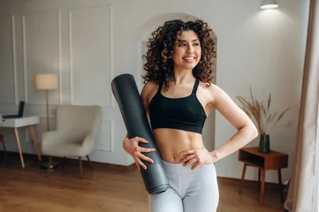 Curly-haired woman in black sports bra and gray leggings holds yoga mat while standing in bright home interior with wooden floor and modern furniture © standret