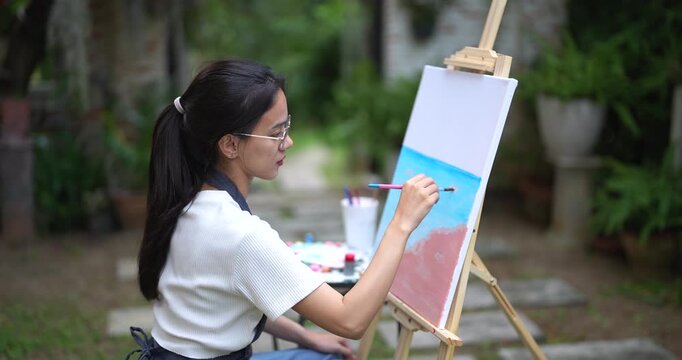 young asian female artist painting sky and sea on canvas during calm creative session sitting on chair in backyard surrounded by potted plants expressing concentration through brush strokes