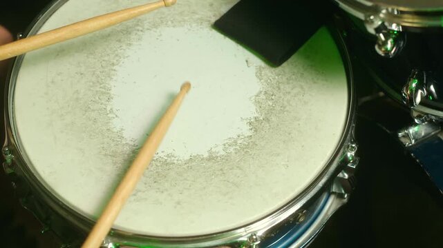 Close up top view musician playing rhythmic beat on snare drum with wooden drumsticks, creating dynamic performance in studio with green stage lighting and dark background. Man playing on snare drum