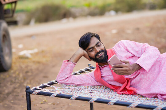 A rural man in traditional attire relaxes on a charpai while using his smartphone