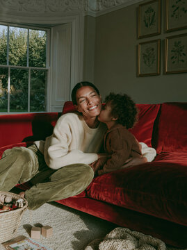 Happy Mother and Child Laughing Together on Red Couch in Cozy Sunlit Living Room With Warm Home Interior and Natural Window Light