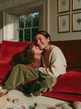 Happy Mother and Child Laughing Together on Red Couch in Cozy Sunlit Living Room With Warm Home Interior and Natural Window Light