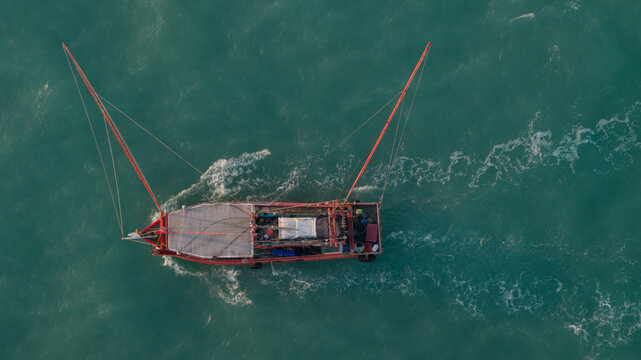 Aerial view of a traditional fishing boat with long red outriggers cruising through turquoise sea water with a white wake Laem Chabang, Chon Buri, Thailand.