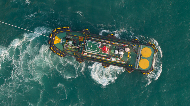 Aerial view of a powerful tugboat navigating through turquoise sea water with a visible tow line and white foam wake in Laem Chabang, Chon Buri, Thailand.
