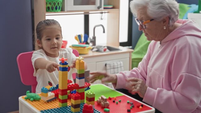 Woman stacking toy blocks on table and pointing finger beside little girl in building; intergenerational bonding joy.
