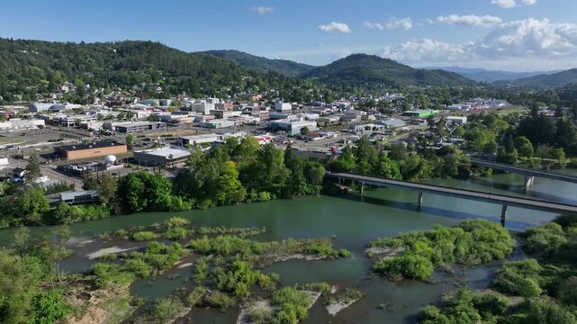 Roseburg downtown along green river with highway bridge, commercial buildings and tree-lined streets tucked among densely forested hills under sunny sky with clouds. Oregon, USA. Aerial wide shot.