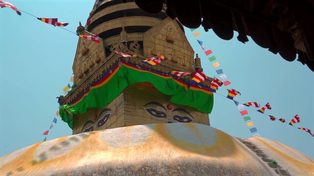 Low angle tracking shot of the golden spire and iconic wisdom eyes of Swayambhunath Stupa decorated with colorful Buddhist flags waving under a blue sky in Kathmandu, Nepal.