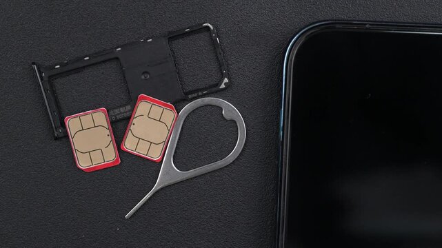 Two SIM cards with red borders, a SIM tray, and an ejector tool are arranged beside a smartphone on a black textured surface. Useful for technology-focused content.
