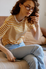 Obraz premium Young woman in a striped shirt sits on a cozy home sofa, casually scrolling on her phone with a thoughtful, relaxed mood under soft natural light in a simple everyday scene.