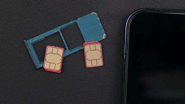 Two red-edged SIM cards and a blue SIM card tray are placed beside a smartphone on a dark surface. Useful for tech reviews or mobile device tutorials.