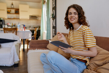 Fototapeta premium A young woman with wavy hair sits on a sofa in a bright living room, reading a notebook and smiling softly, creating a warm, cozy home lifestyle moment for audiences.